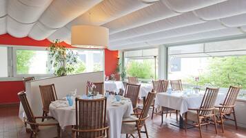 Un restaurante con varias mesas, sillas de mimbre, ventanas amplias y un techo blanco.