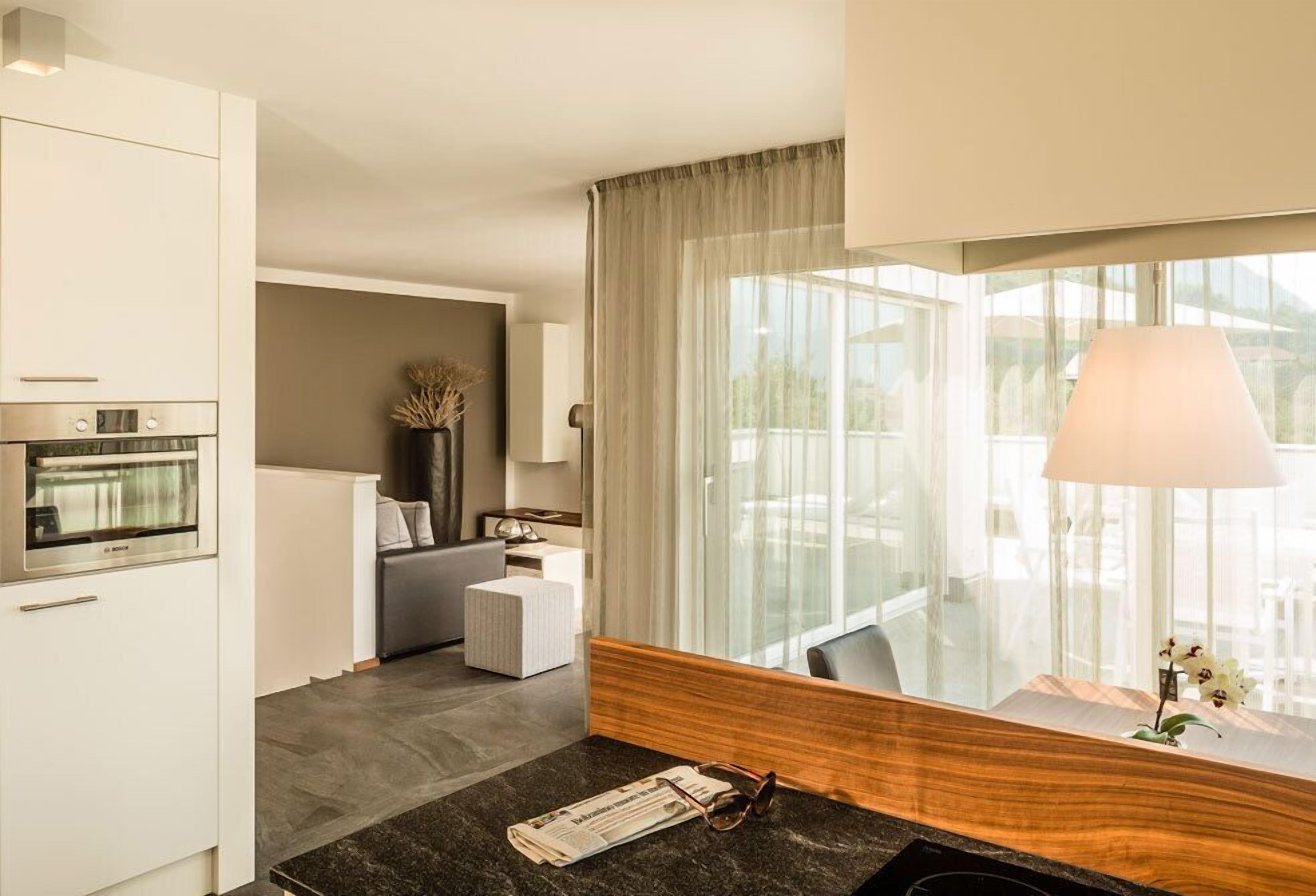 Una habitación de hotel moderna con área de cocina, zona de estar con sofá y una pequeña mesa, y un amplio ventanal con cortinas.