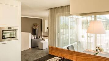 Una habitación de hotel moderna con área de cocina, zona de estar con sofá y una pequeña mesa, y un amplio ventanal con cortinas.