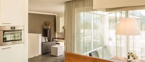 A modern hotel room with a kitchen area, a seating area with a sofa and a small table, and a large window with curtains.