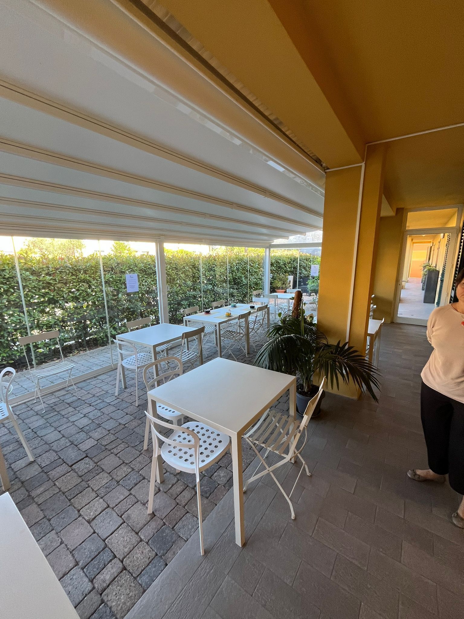 Una terrazza esterna con tavoli e sedie, una pianta in vaso e un vialetto che conduce all'ingresso di un edificio.