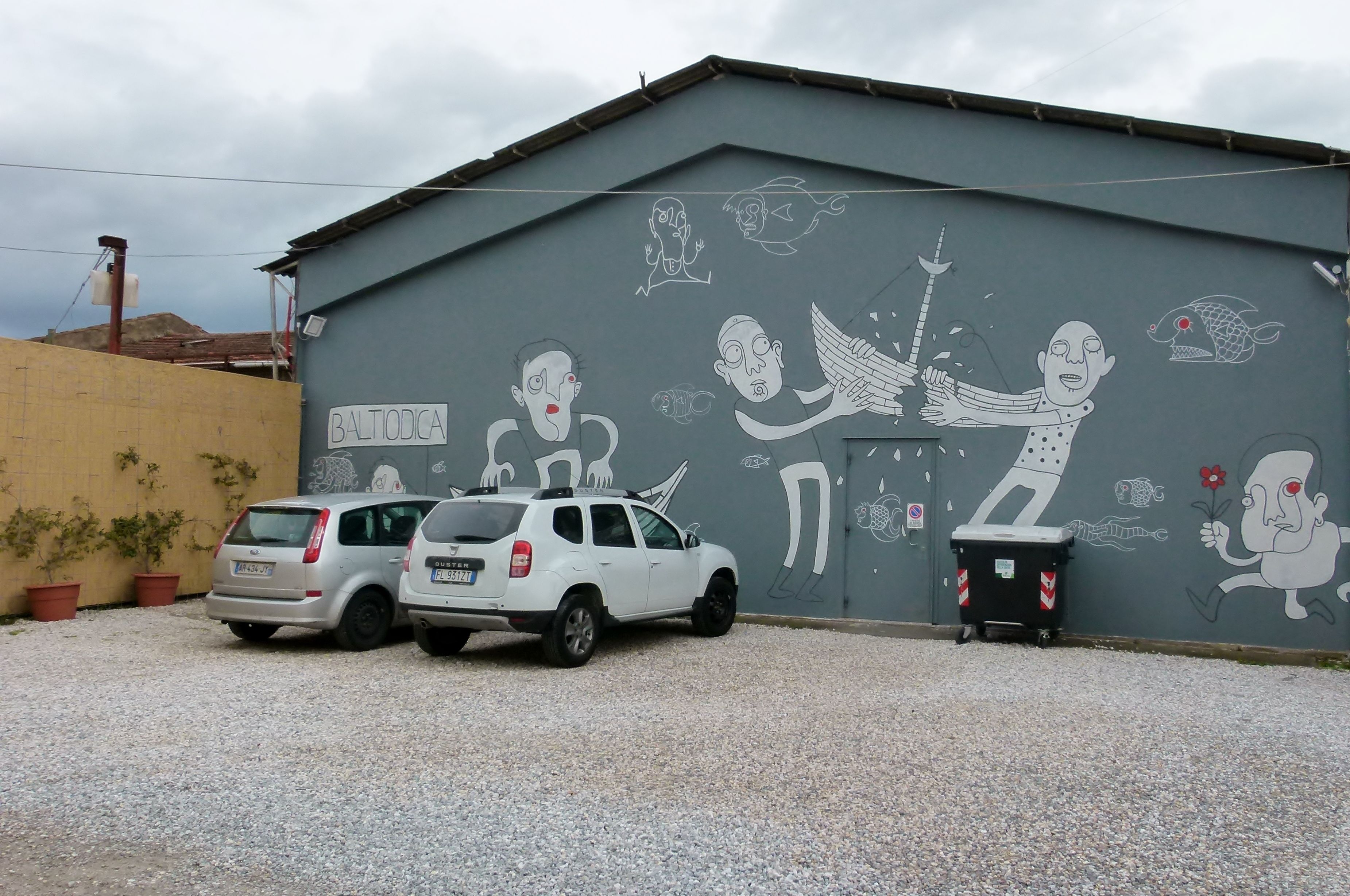 Un bâtiment dont le mur gris est orné d’une fresque représentant divers personnages, et sur lequel on peut lire l’inscription « BALTIODICA ».