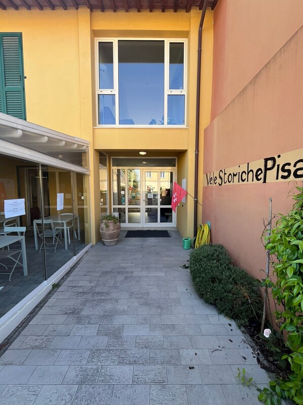 A building with a yellow facade, a glass door entrance, outdoor seating, and a sign reading 'Vele Storiche Pisa'. - Vele Storiche Pisane  (Pisa)