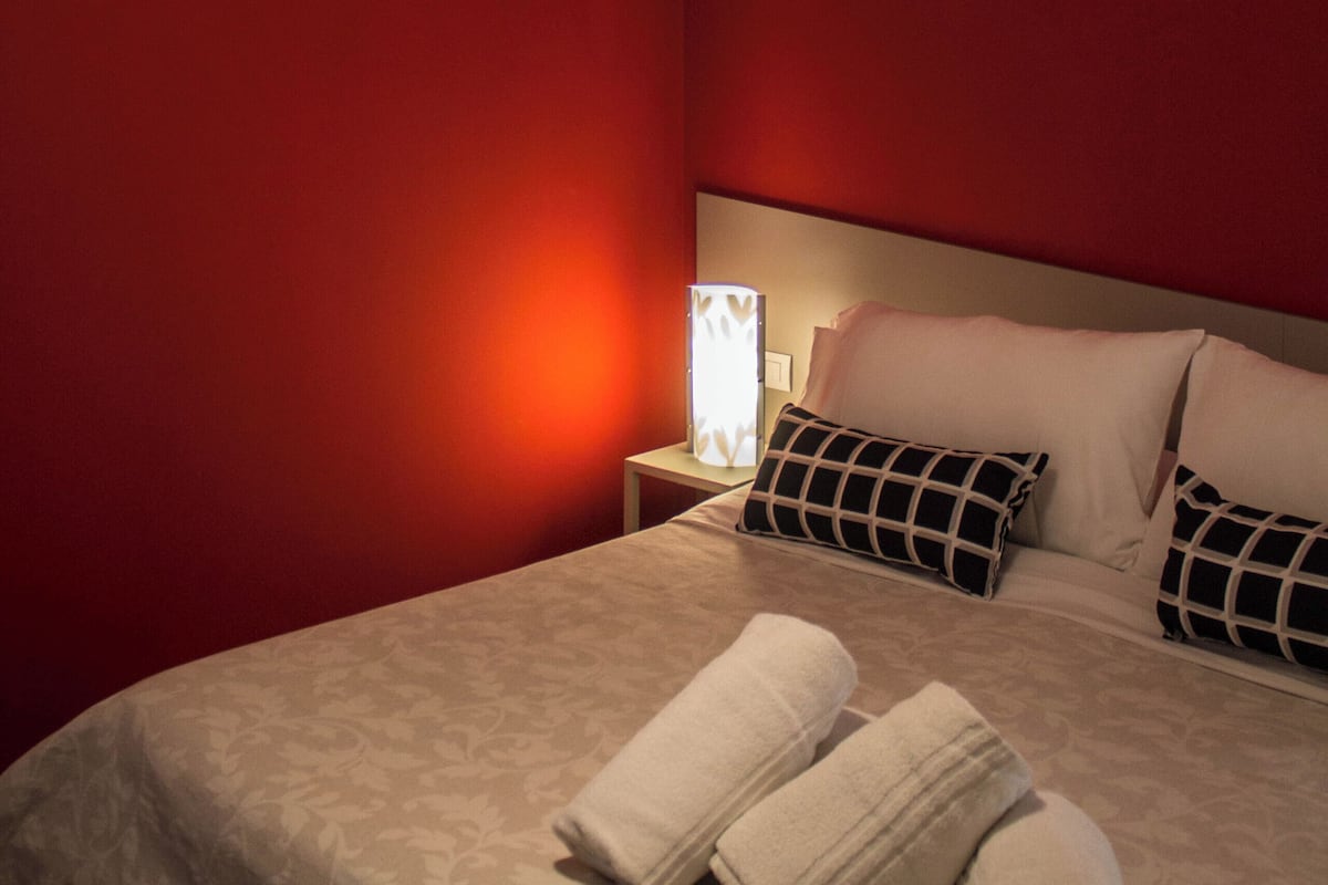 A hotel room with a red wall, a bed with a beige bedspread, two pillows, a nightstand with a lamp, and two rolled towels on the bed.