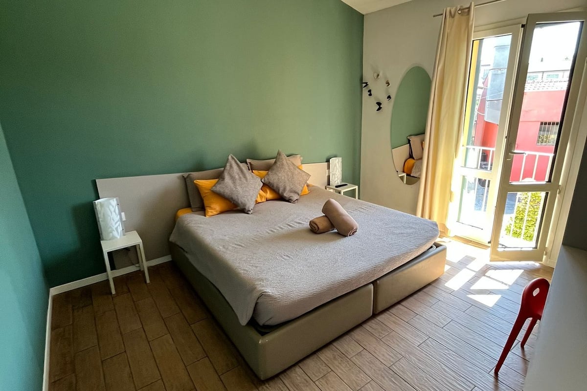 A bedroom with a bed, a small bedside table, a red chair, and a view of the outside.