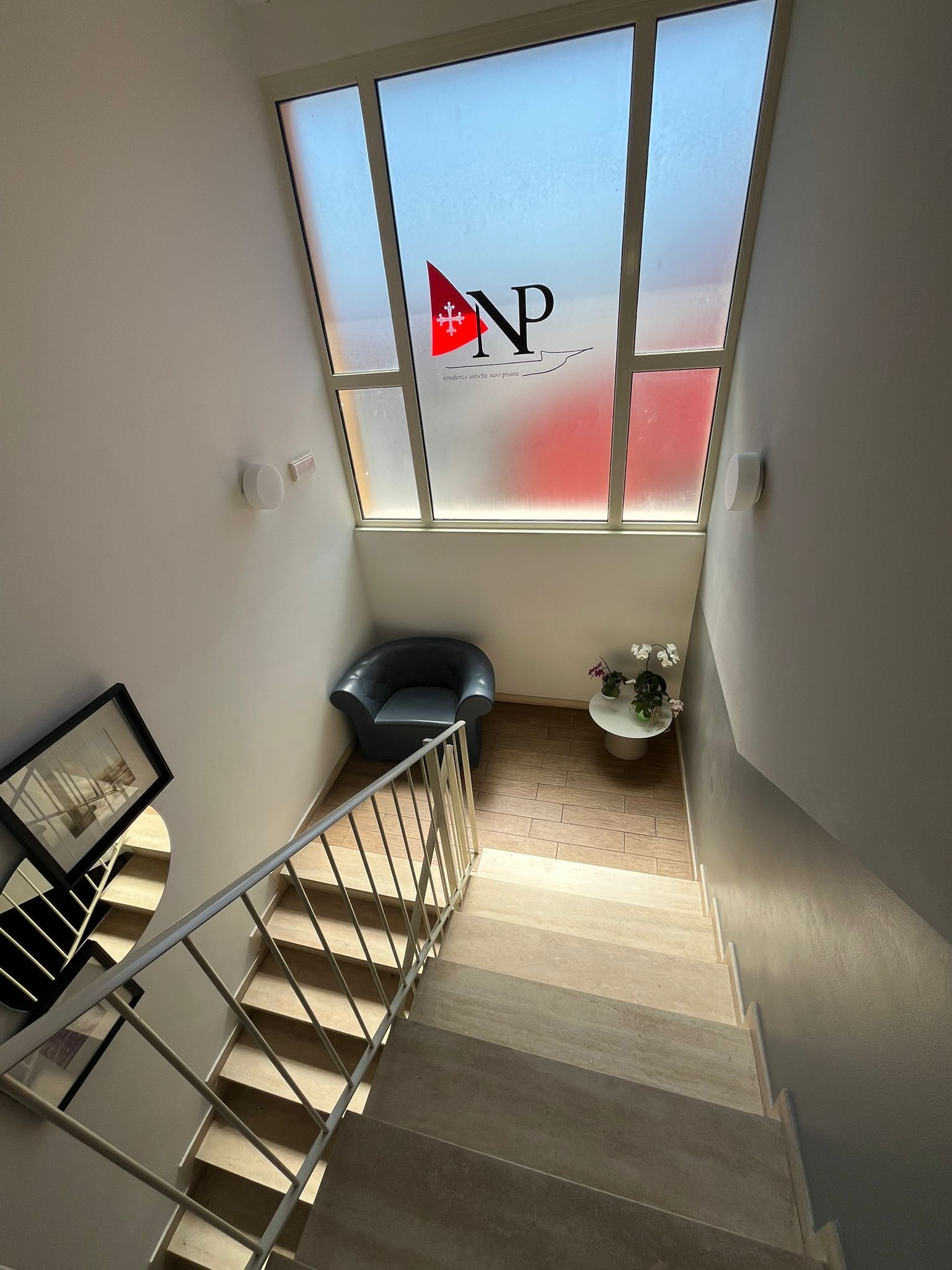 Un escalier mène à une pièce dotée d’une fenêtre sur laquelle est affiché un logo et un drapeau. On y trouve également une chaise, une petite table avec une plante et un tableau encadré accroché au mur.