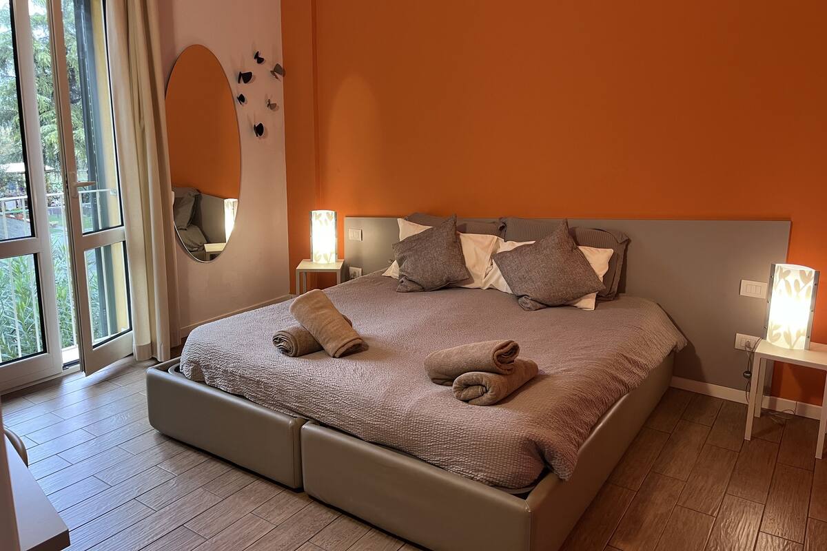 A bedroom with a large bed, two bedside tables, a mirror, and a view of greenery outside.