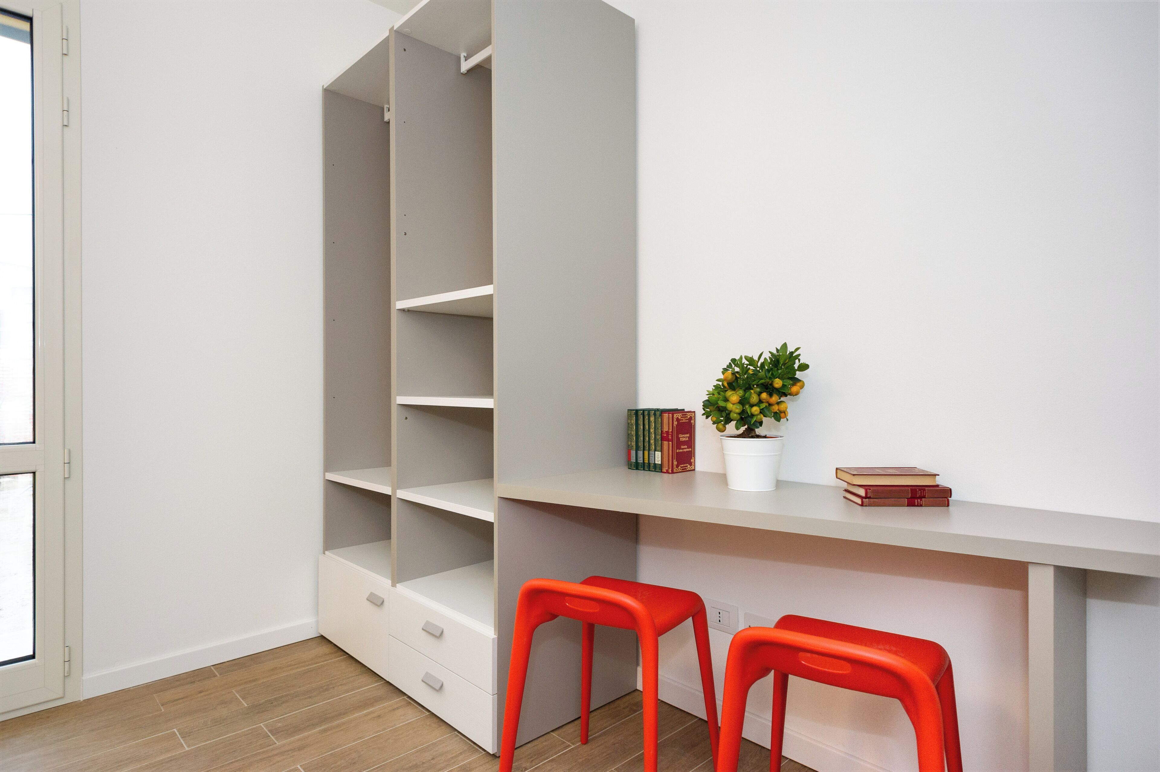 A room with a white desk, two red stools, a potted plant, and a bookshelf.