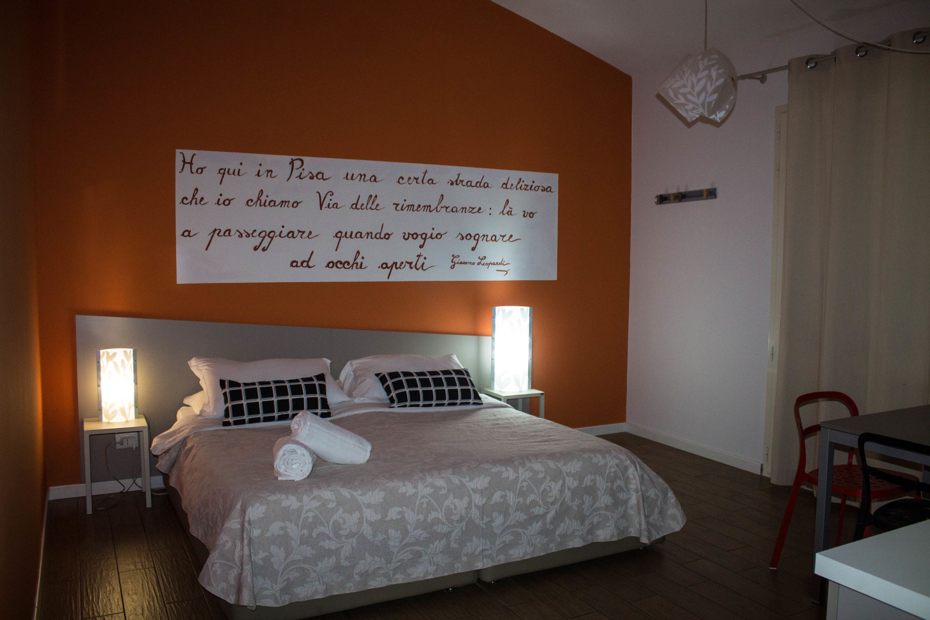 Ein ordentlich gemachtes Bett mit einem gemusterten Bettbezug und Kissen, einer Nachttischlampe sowie einer Wand mit einem Zitat auf Italienisch.