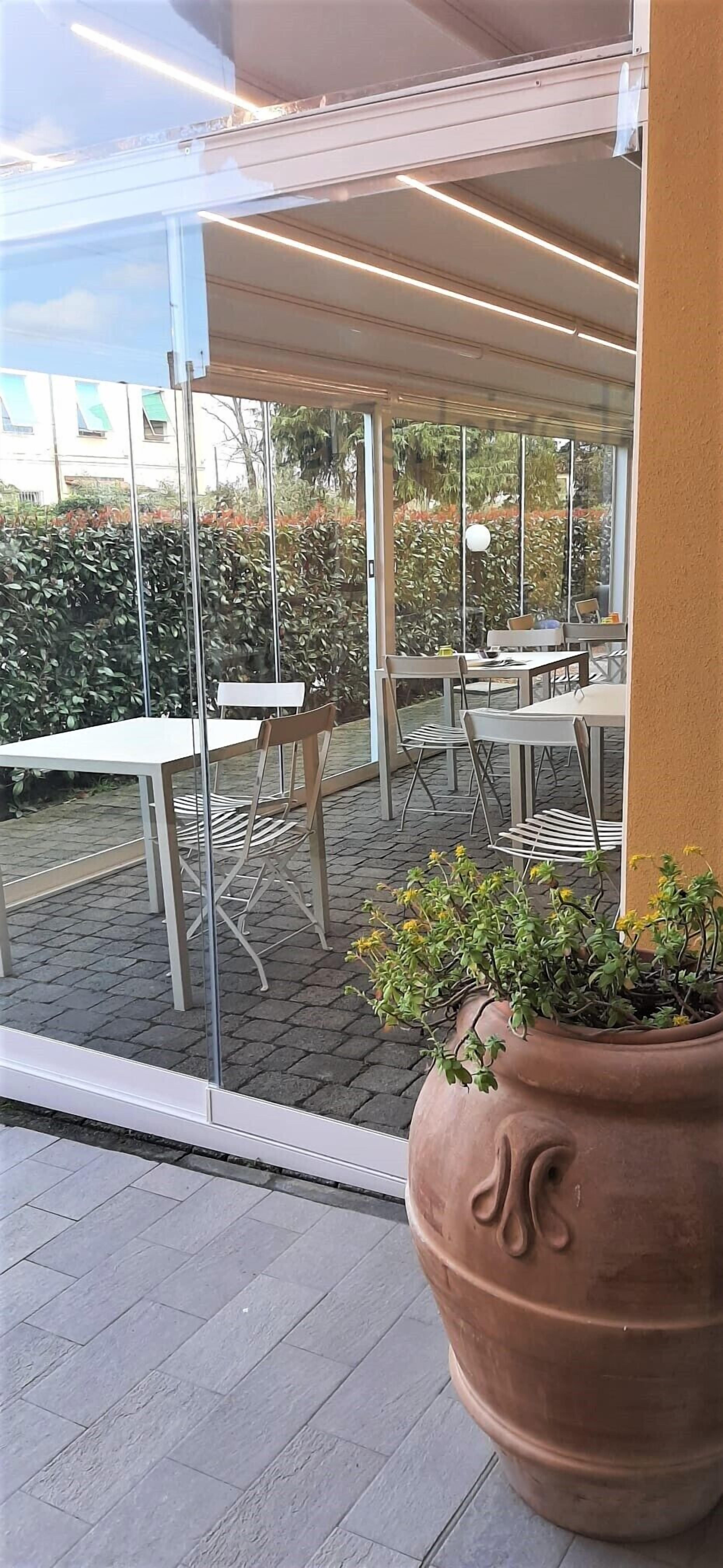 En udendørs patio med glasvægge, borde, stole og en stor terracotta-potte med planter.