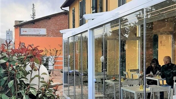 Un café al aire libre con mesas y sillas, un cerramiento de vidrio y un edificio con paredes amarillas y contraventanas verdes al fondo.