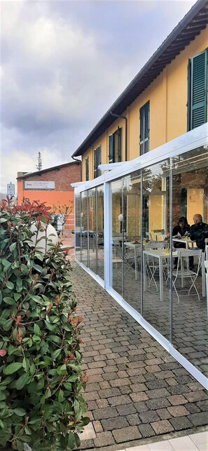 An outdoor cafe with tables and chairs, a glass enclosure, and a building with yellow walls and green shutters in the background.