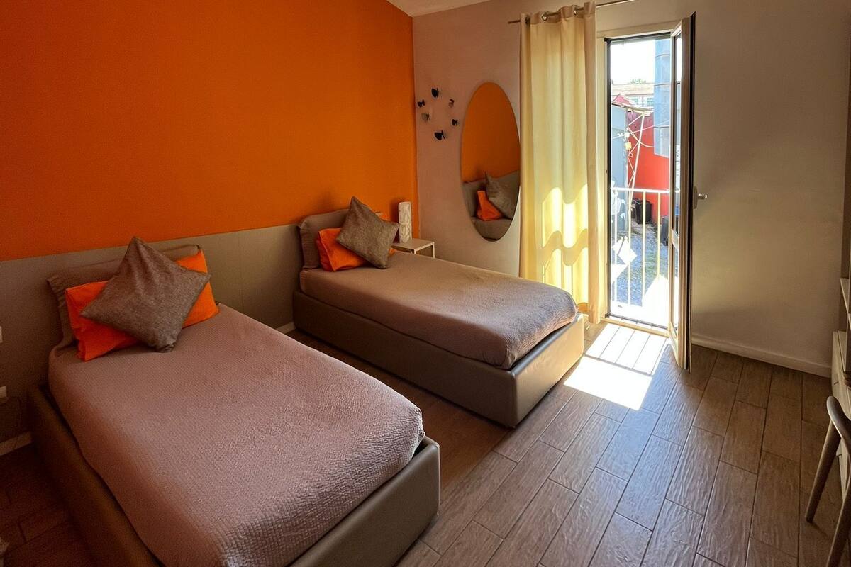 A room with two beds, a window with curtains, a mirror, and a view of the outside.