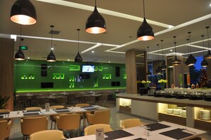 Een modern restaurant met een bar verlicht in groen, meerdere eettafels en hanglampen.
