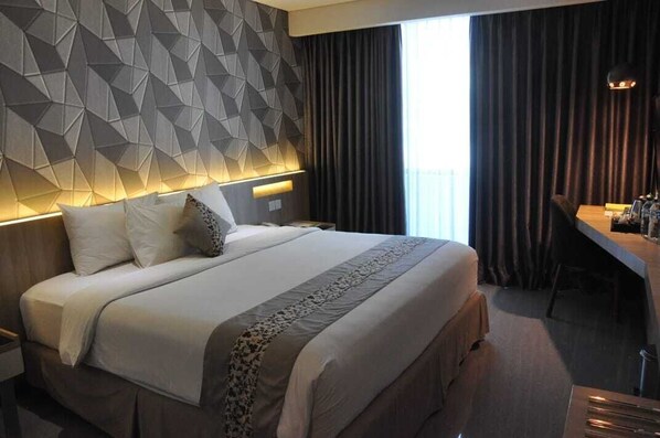 Een moderne hotelkamer met een hoofdeinde met een geometrisch patroon, een groot bed met een bedovertrek met dessin, een bureau met stoel en uitzicht op het groen buiten het raam.