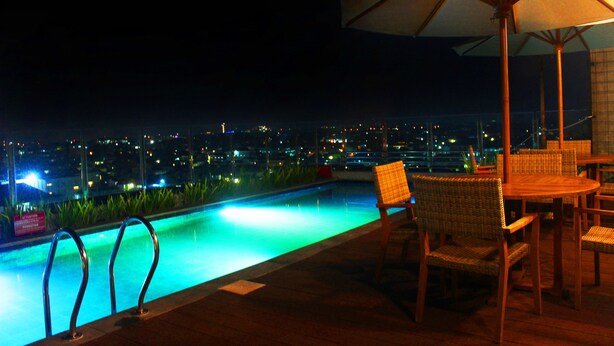 Zona de piscina nocturna con vistas a las luces de la ciudad, una mesa de madera con sillas y una sombrilla.