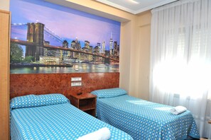 A hotel room with two single beds, a cityscape mural on the wall, a wooden headboard, and a window with sheer curtains.