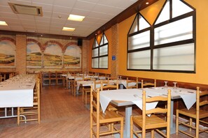 A dining area with long tables and chairs, a mural on the wall, and large windows.