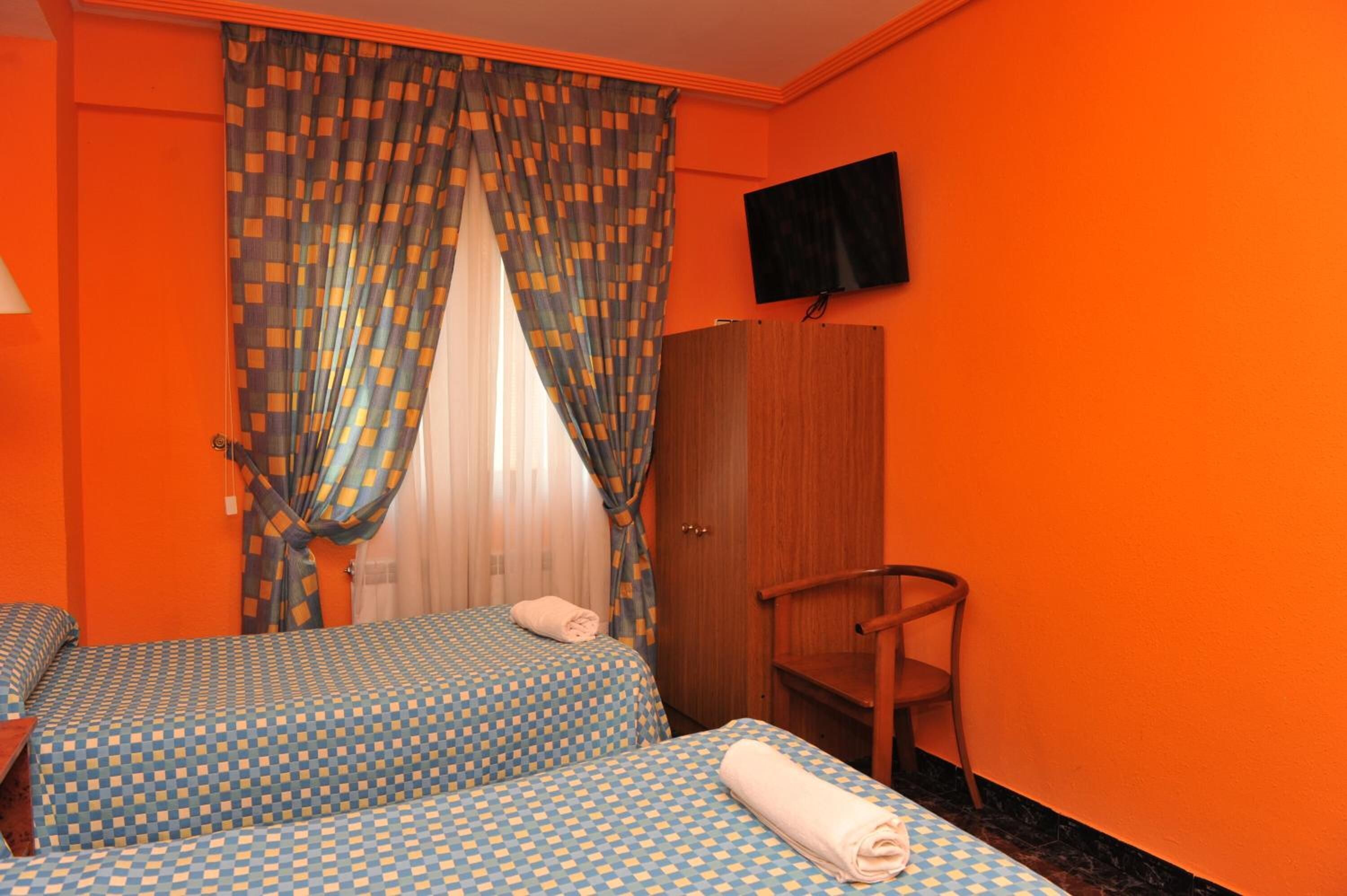 A room with two beds, a wooden wardrobe, a television on the wall, and a window with patterned curtains.