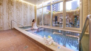 Zona d'spa amb una piscina envuallada amb vidre, una dona asseguda al costat i vista exterior a través de grans finestres.