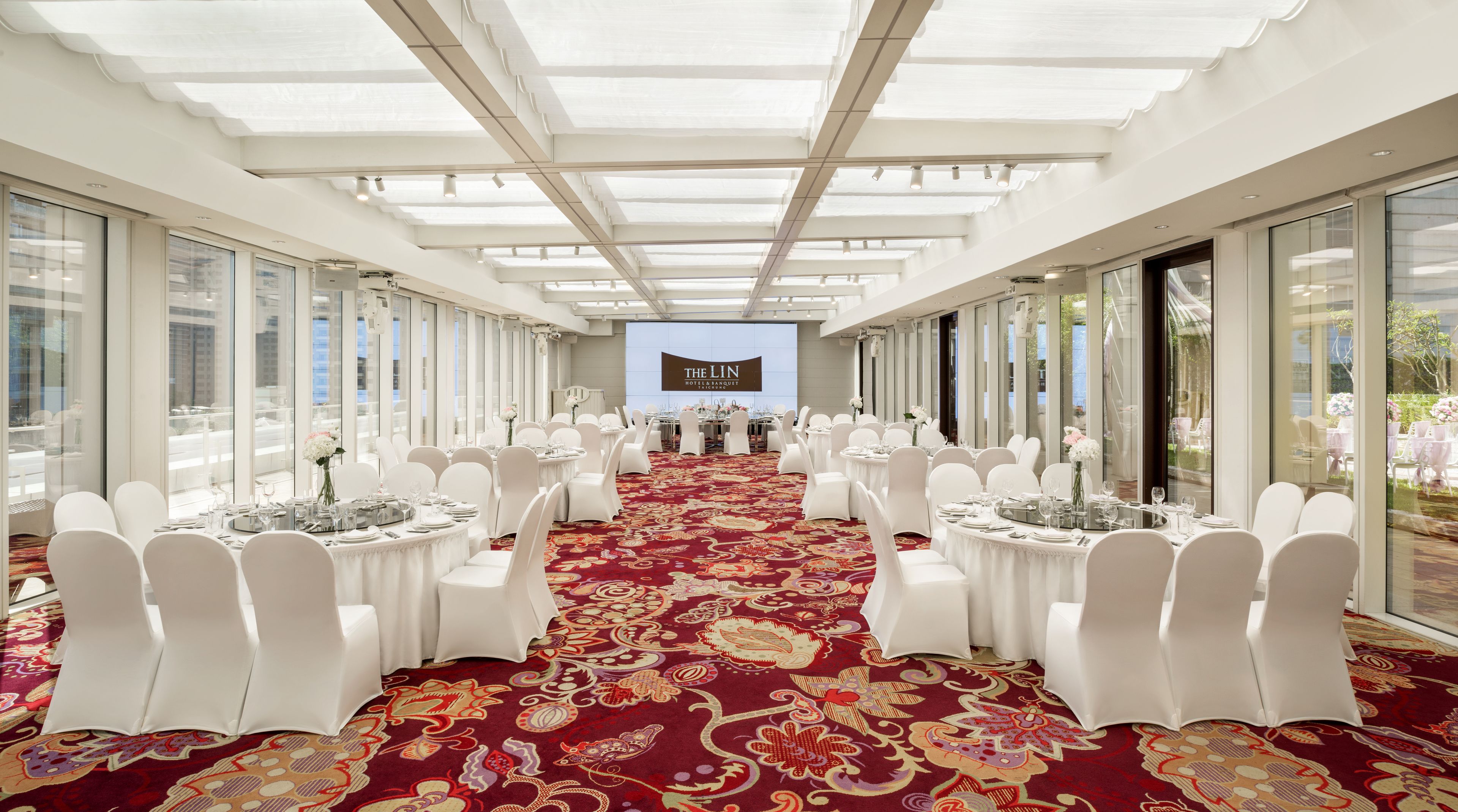A banquet hall with white chairs and tables, a red carpet, and a large screen at the front.