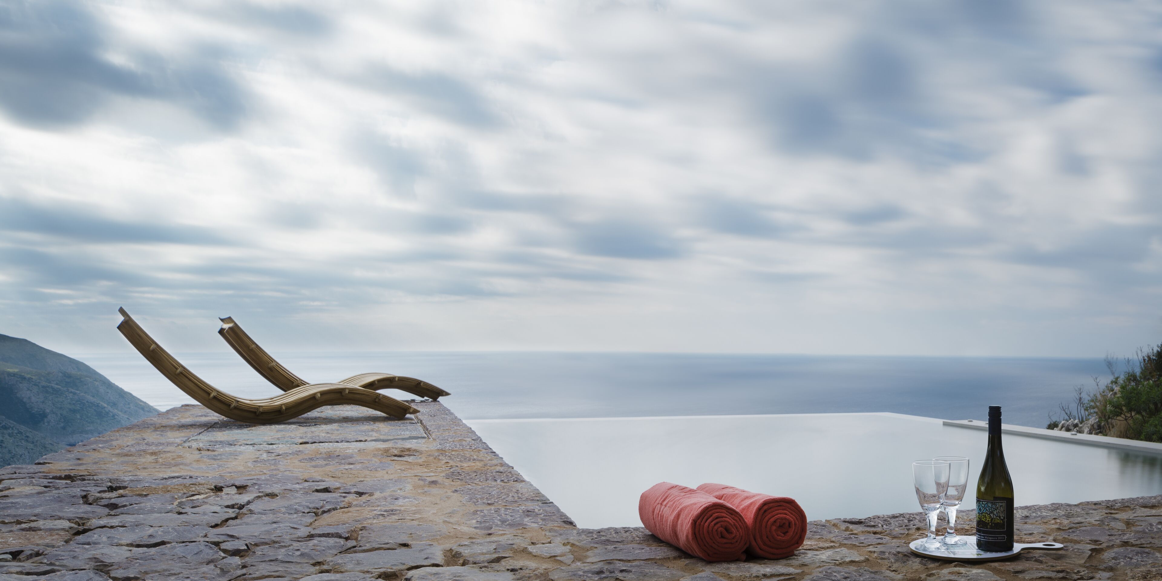 Un saliente de piedra con una botella de vino, copas y una toalla enrollada, con vistas a una masa de agua tranquila y montañas lejanas.