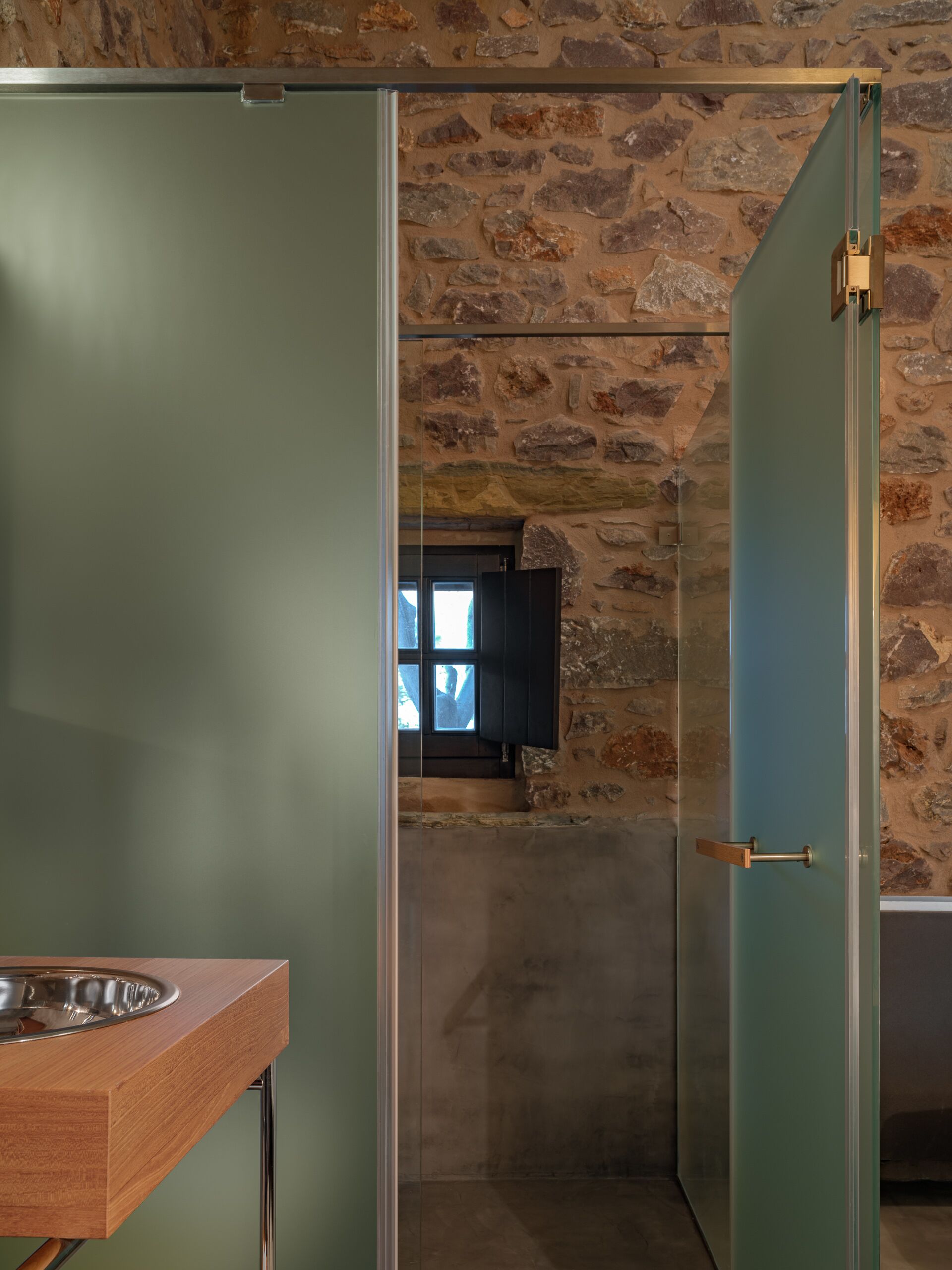 Baño con pared de piedra, lavabo de madera y puerta de cristal.