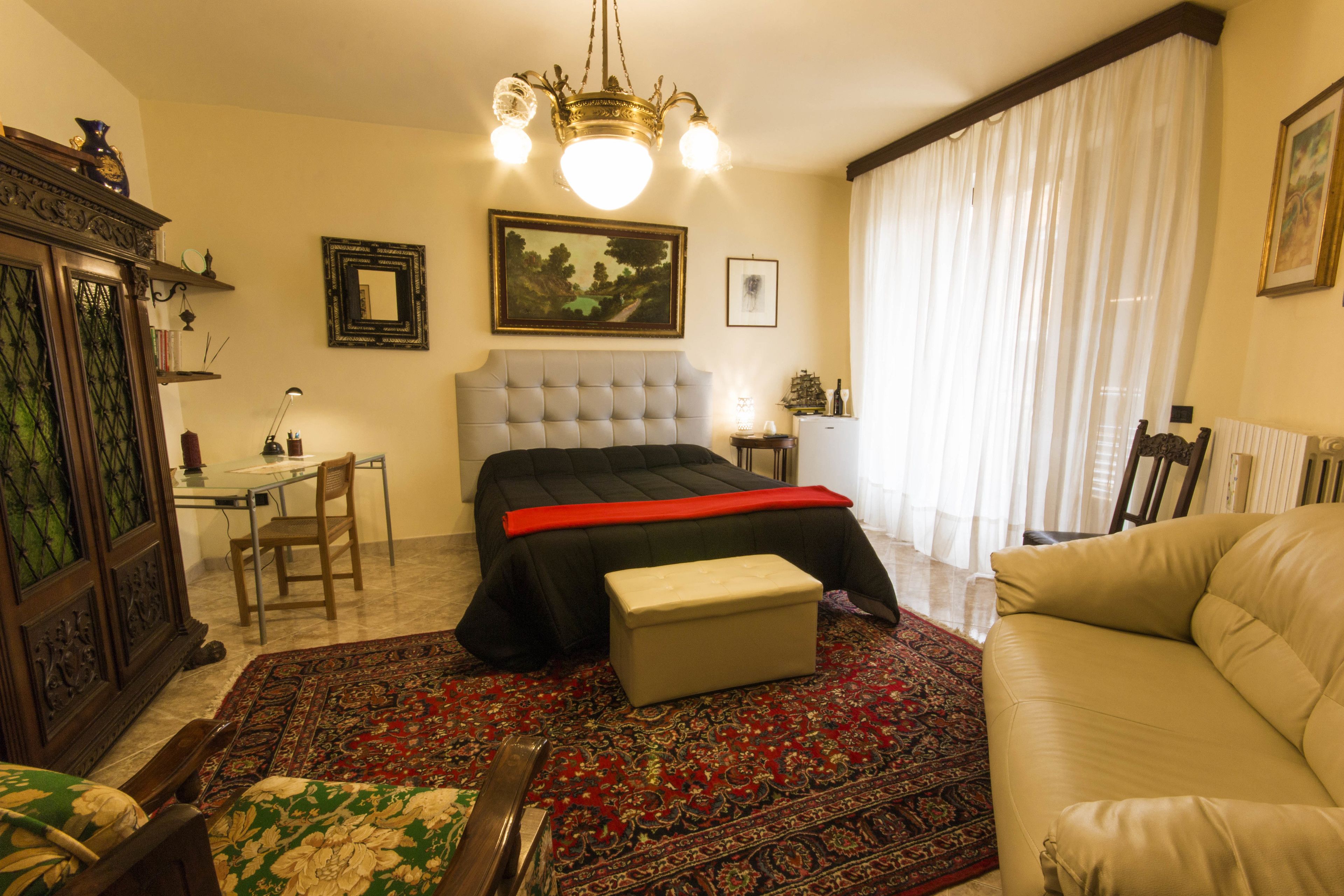 Une chambre d’hôtel comprenant un lit, un canapé, un bureau avec une chaise, un lustre et une grande fenêtre avec des rideaux.