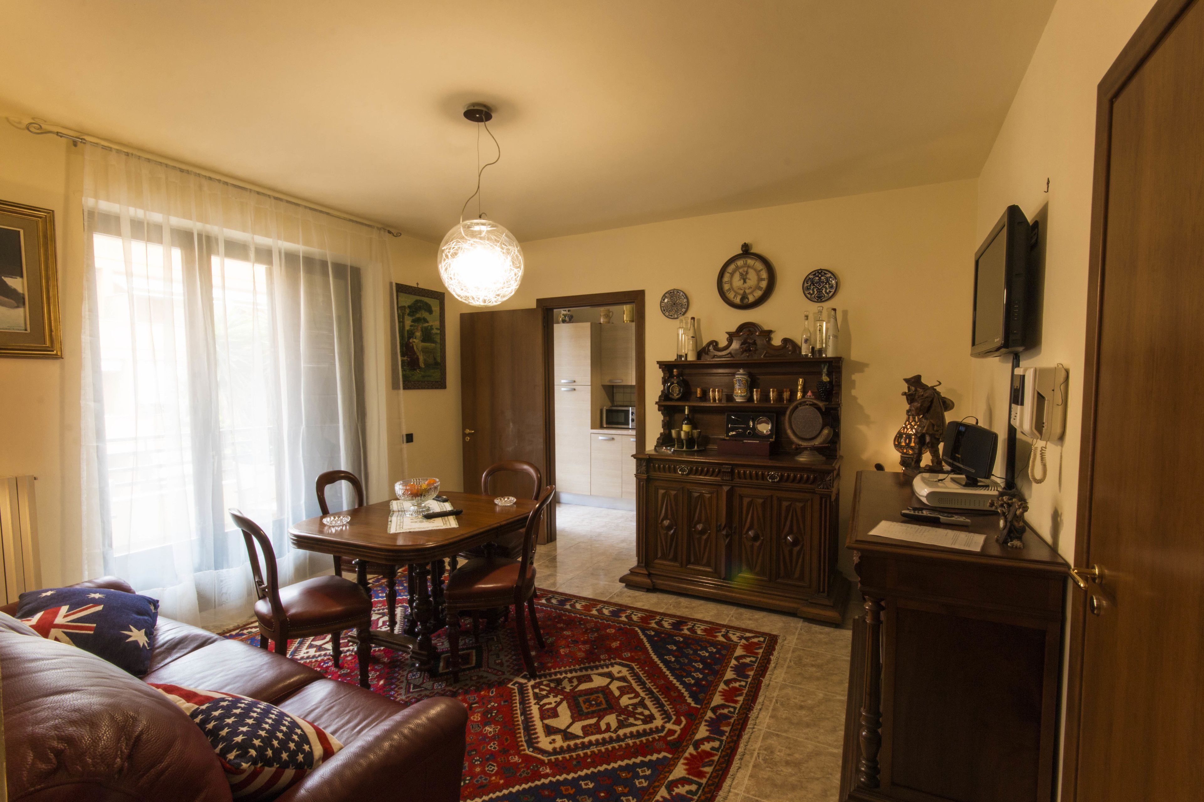 A living room with a dining table, chairs, a sofa, a patterned rug, a wooden cabinet, a television, and a clock on the wall.