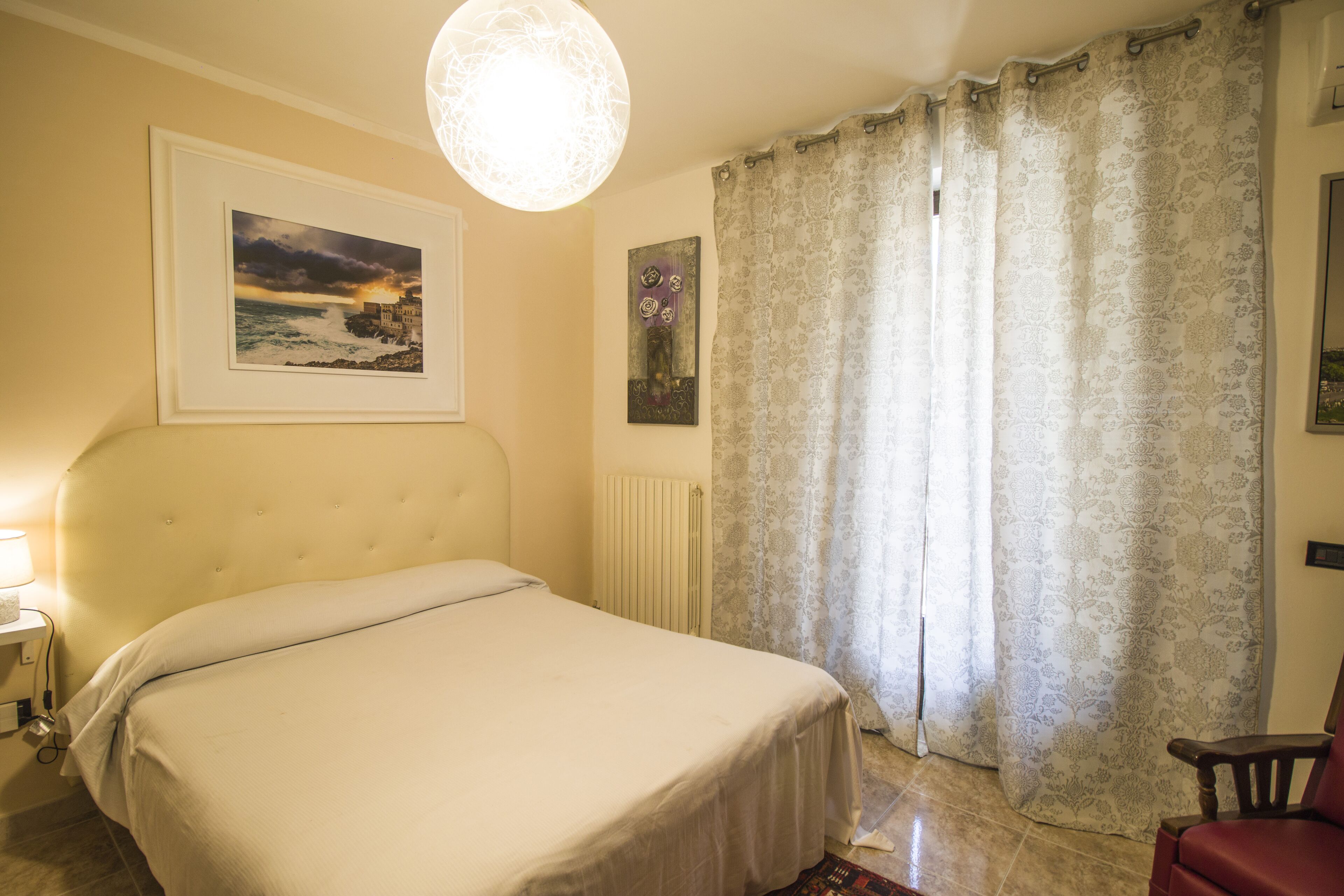 A bedroom with a large bed, a radiator, a framed picture of a coastal scene, and a window with patterned curtains.