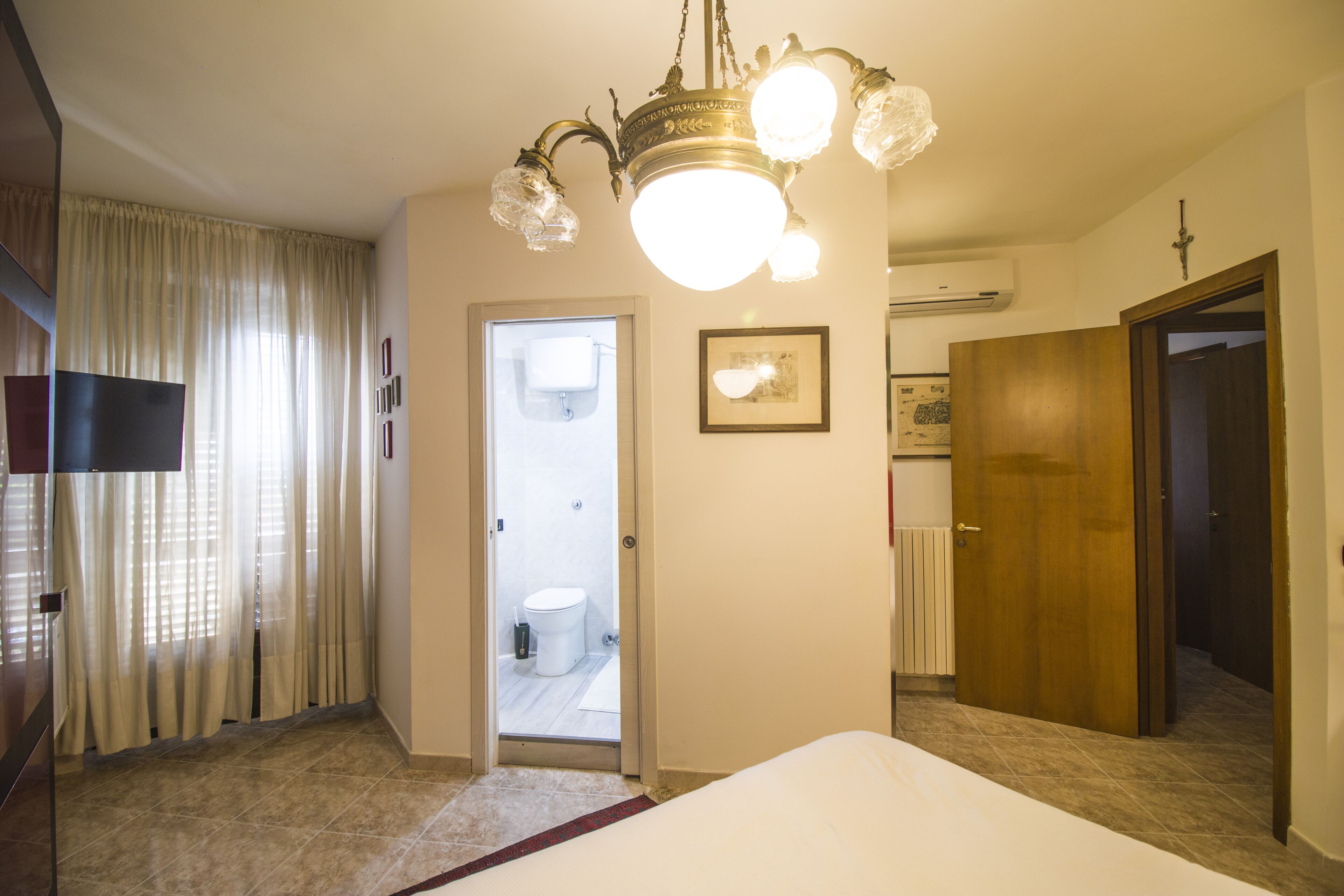 A hotel room with a bed, a chandelier, a door leading to a bathroom, and a television on the left.