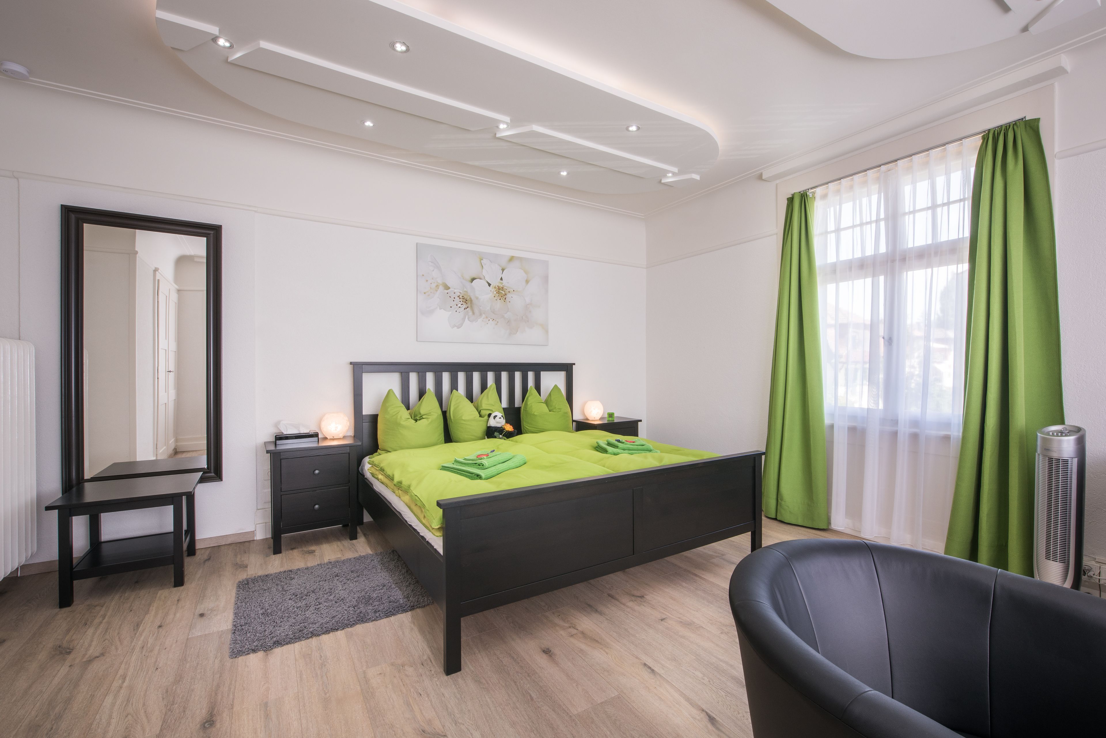 A modern bedroom with a black bed, green bedding, a bedside table, a mirror, a nightstand, a lamp, a chair, and a window with green curtains.