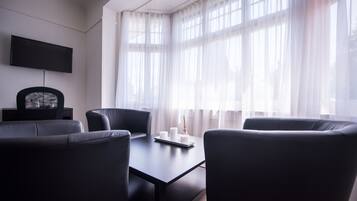 A modern room with a dark-colored sofa set, a flat-screen TV mounted on the wall, and a large window with sheer curtains.