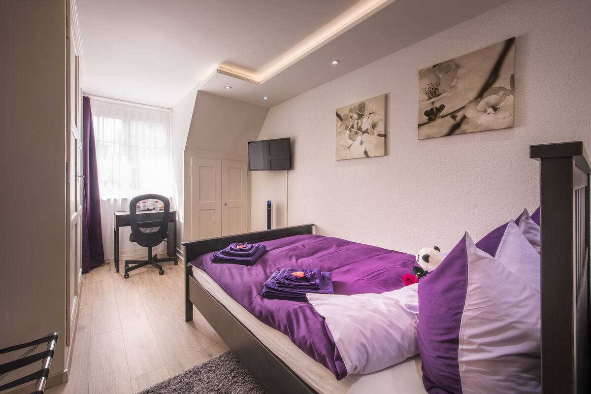 A bedroom with a bed, a desk, a chair, a TV, and artwork on the wall.