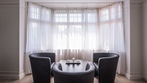 Uma sala de jantar moderna com uma mesa redonda e duas cadeiras pretas, grandes janelas com cortinas leves e piso de madeira.