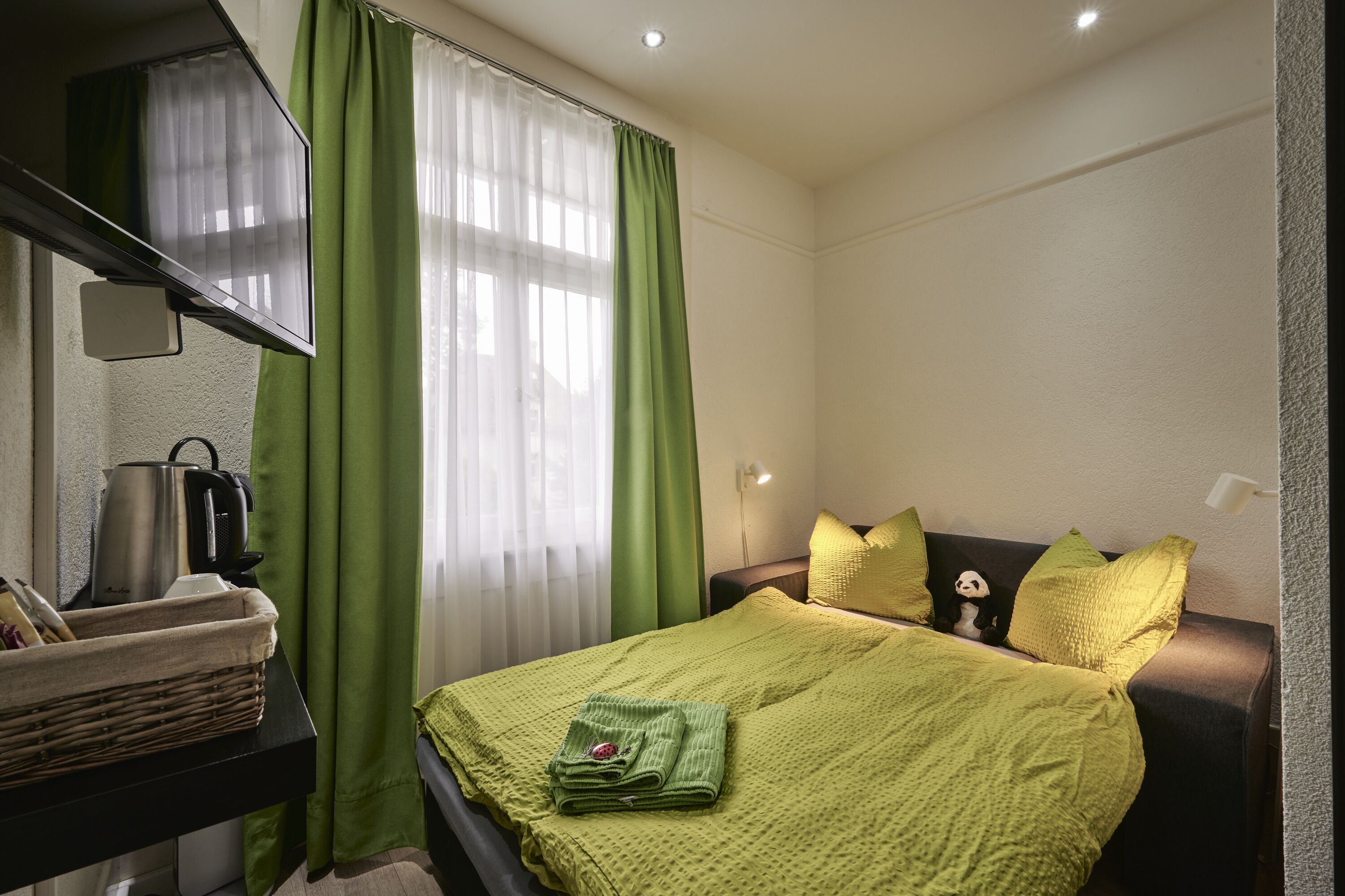 Un lit bien fait, recouvert d’une couverture verte, une table de chevet avec une bouilloire et un rideau vert.
