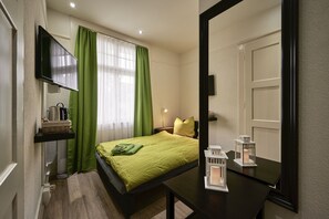 Un dormitorio con cama, televisor montado en la pared, mesita de noche con lámpara y ventana con cortinas verdes.