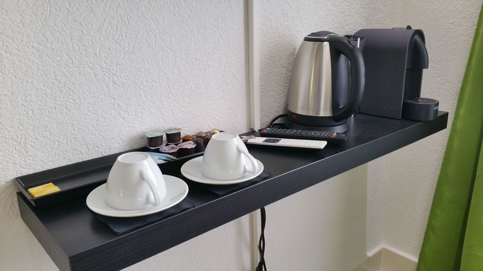 Ein schwarzes Tablett mit zwei weißen Tassen und Untertellern, einem silbernen Wasserkocher, einer schwarzen Kaffeemaschine und einer kleinen Schale mit verschiedenen Pralinen vor einer weißen Wand.