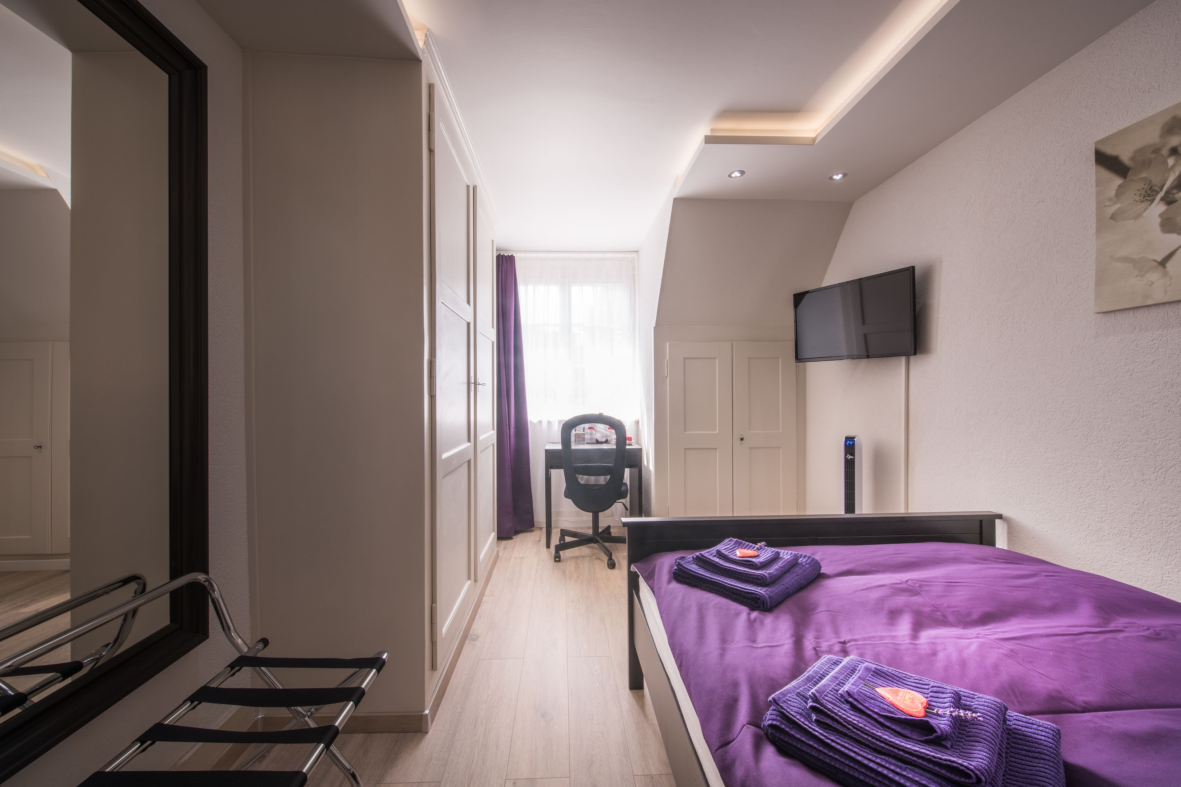 A bedroom with a bed, a desk, a chair, a TV, and a mirror.