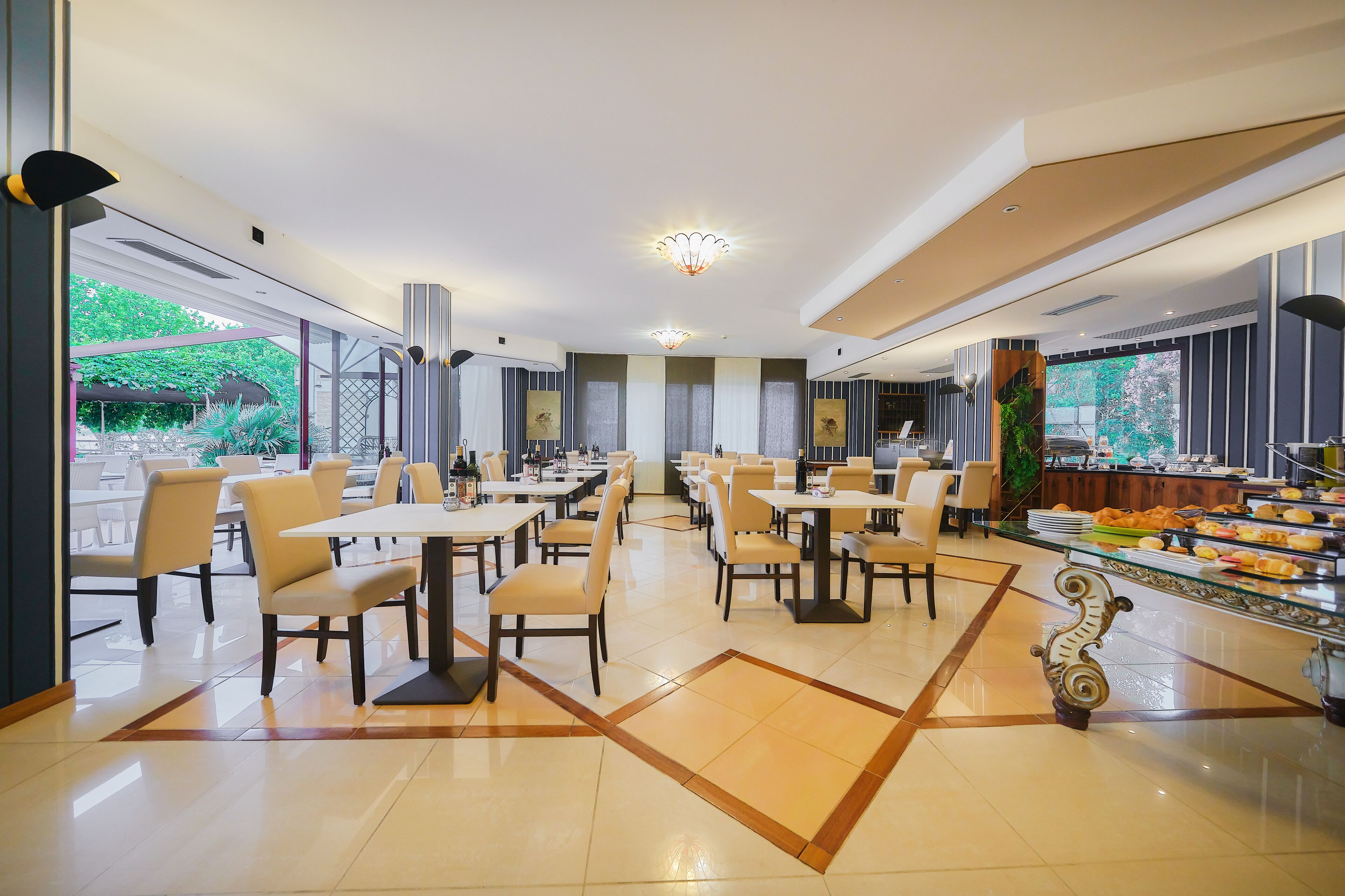 A modern dining area with tables and chairs, a buffet with various food items, and large windows offering a view of greenery outside.