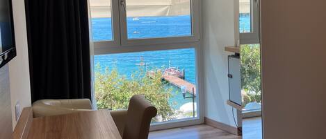 A hotel room with a wooden desk, a chair, a bed, a TV, and a large window with a view of the sea and a dock.