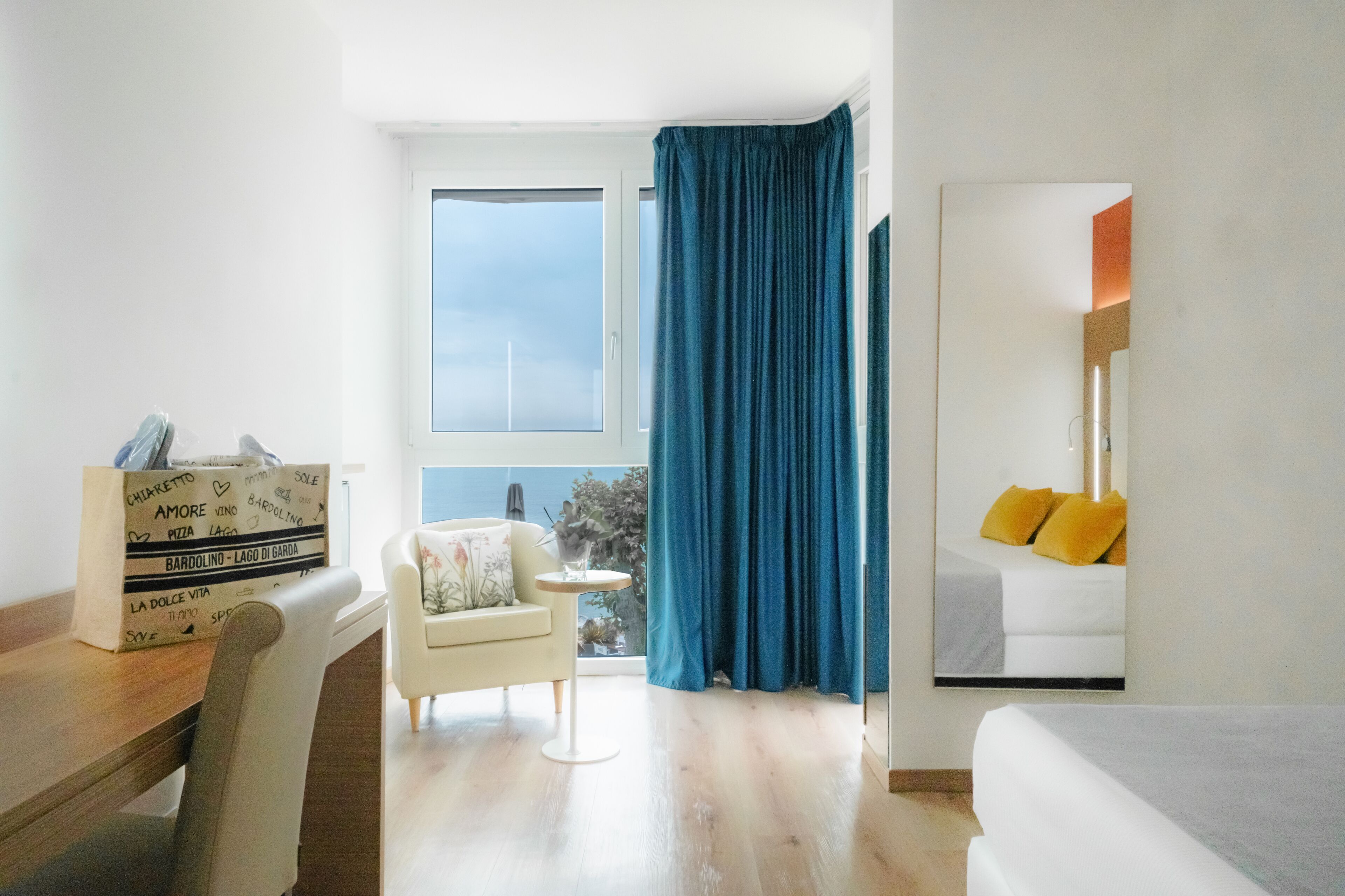 Une chambre d’hôtel avec une grande fenêtre, un lit avec des oreillers jaunes, une petite table ronde avec une chaise, et un bureau en bois sur lequel se trouve une boîte à pizza.