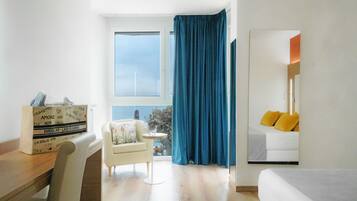 A hotel room with a large window, a bed with yellow pillows, a small round table with a chair, and a wooden desk with a box of pizza.