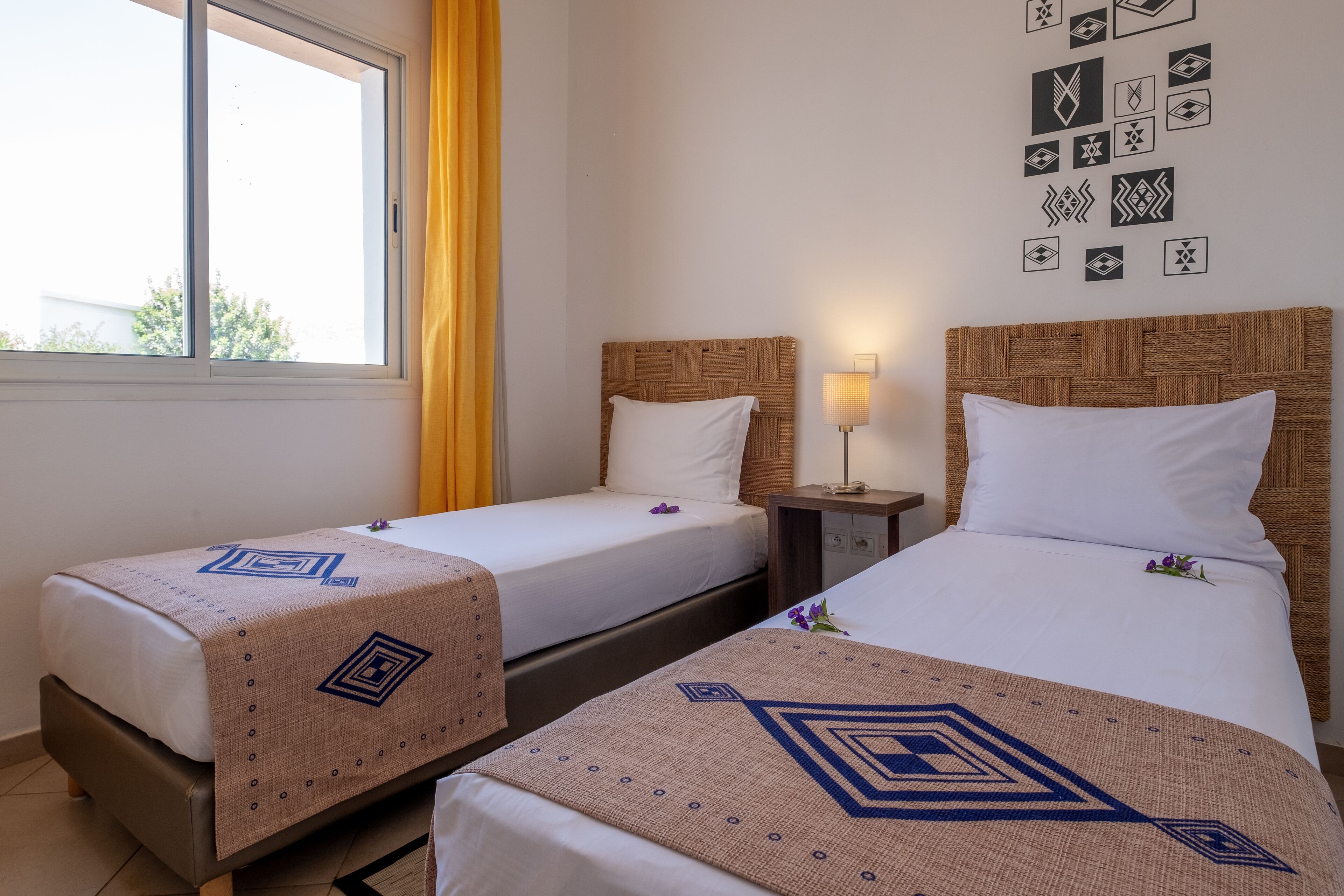 Una camera d'albergo con due letti, una finestra con tende gialle e un'opera d'arte geometrica sulla parete.