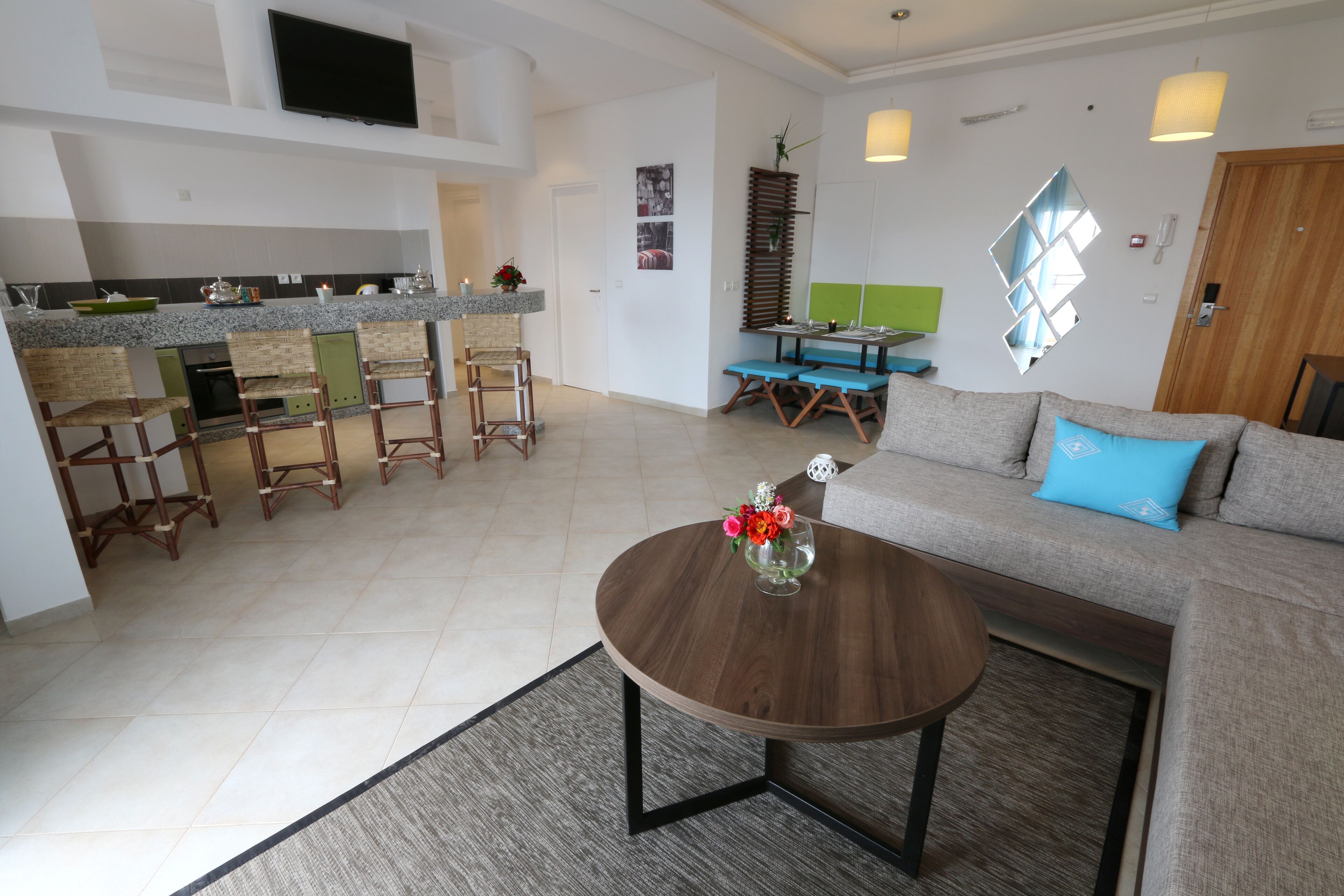 A modern living room with a sofa, coffee table, and bar stools. A kitchen with a TV and counter is visible in the background.