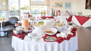 A buffet setup with various food items and serving utensils on a table, set in a room with a view of the outdoors. - Lunja Village Taghazout (Tamri)