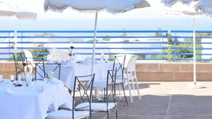 An outdoor dining area with white tables and chairs, white umbrellas, and a view of the sea. - Lunja Village Taghazout (Tamri)