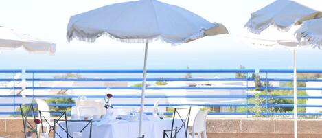 An outdoor dining area with white tables and chairs, white umbrellas, and a view of the sea.