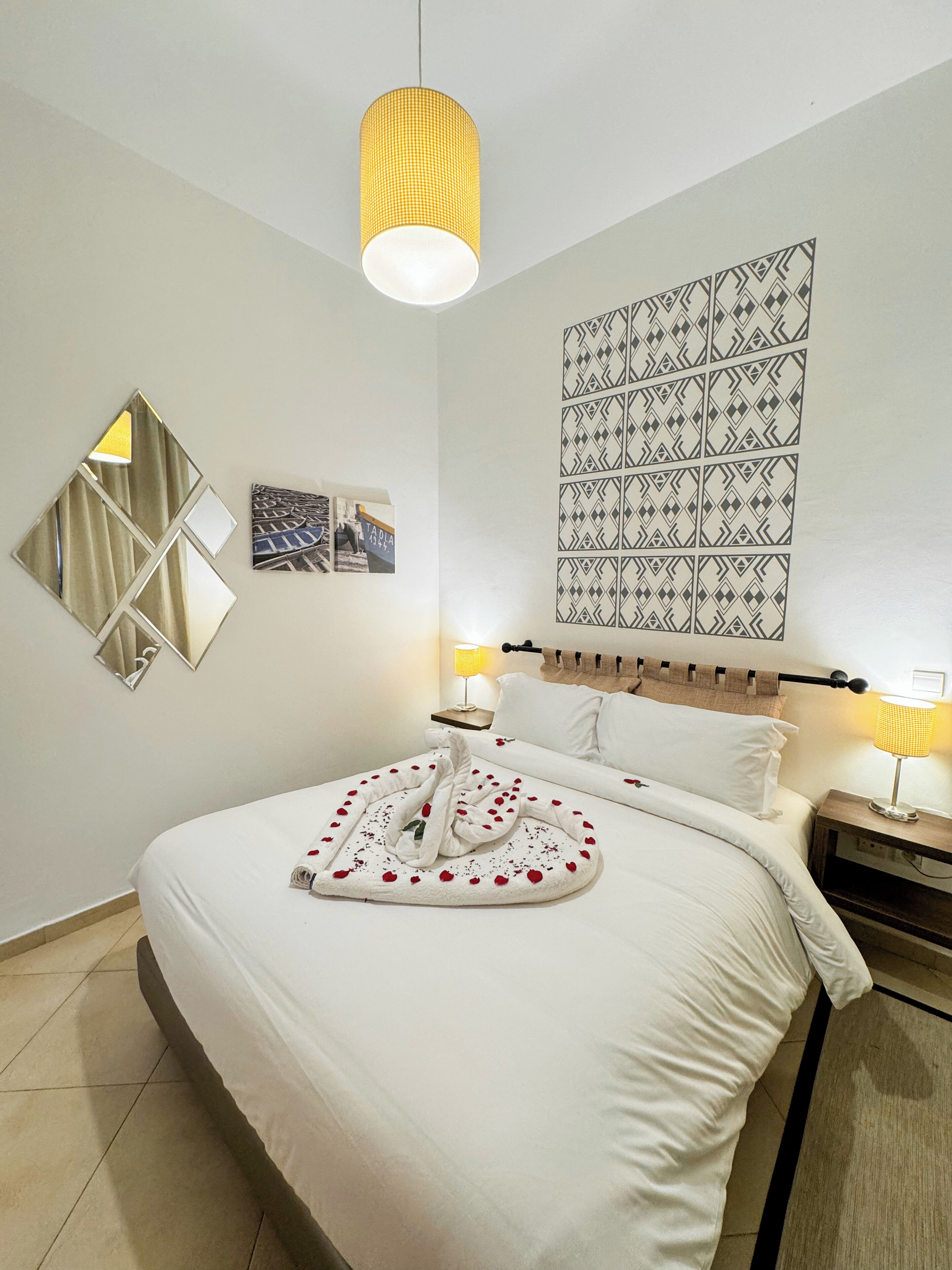 Una cama bien hecha con una toalla y pétalos de rosa, un tapiz de pared estampado y un espejo geométrico en la pared.