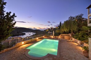 Une piscine dotée d’une terrasse en pierre, entourée de chaises longues et offrant une vue sur la mer et les collines.
